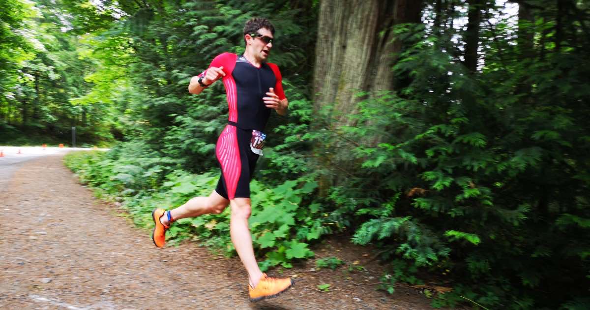 A man in a black and white triathlon body suit running along a forest trail.